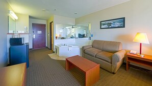 Hotellihuone, jossa on sohva, pieni pöytä, lamppu, televisio, mikroaaltouuni, jääkaappi, kylpyammeella varustettu kylpyhuone ja ovi.