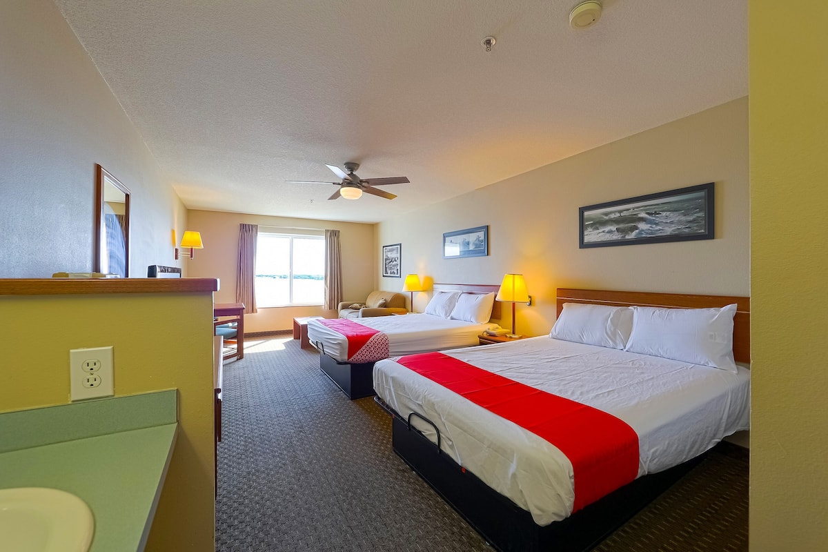 A hotel room with two beds, a ceiling fan, a window with curtains, a desk with a lamp, and a painting on the wall.