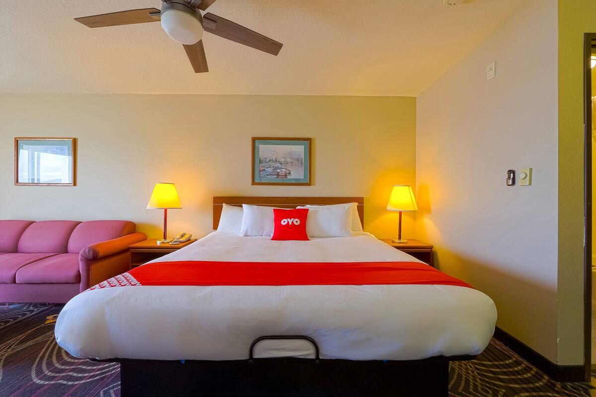 A hotel room with a bed, a desk, a chair, a ceiling fan, two lamps, and a framed picture on the wall.