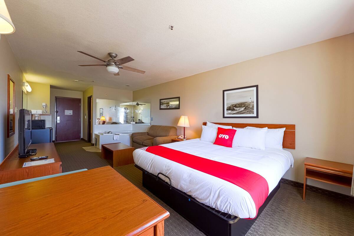 A hotel room with a bed, a desk, a chair, a small table, a ceiling fan, and a picture on the wall.