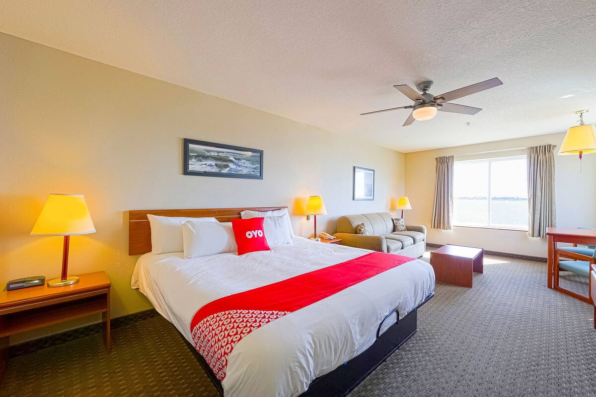 A hotel room with a large bed, a ceiling fan, a desk, a chair, and a window with curtains.