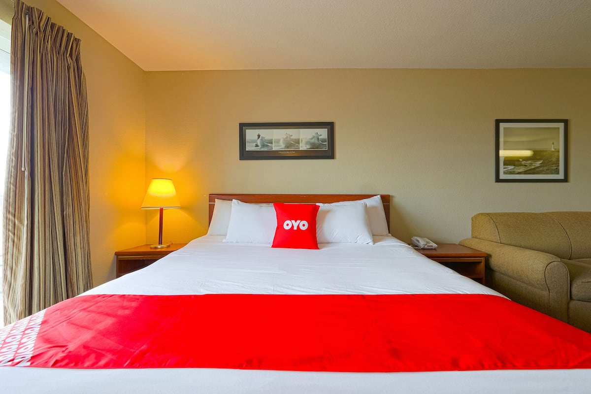 A hotel room with a bed, a nightstand with a lamp, a chair, and two framed pictures on the wall.