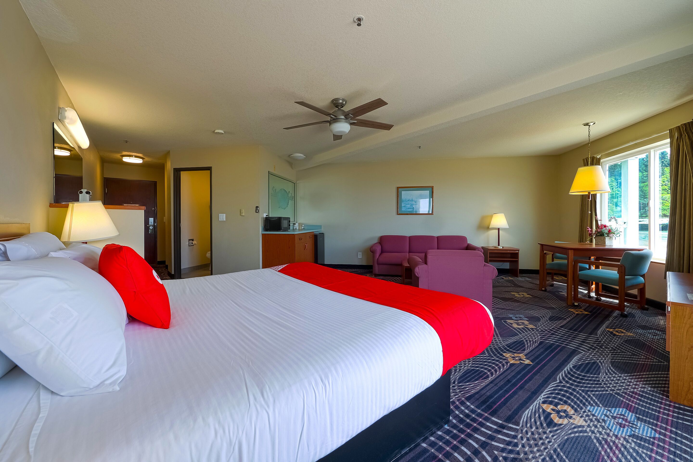 A hotel room with a large bed, a ceiling fan, a desk with a chair, a sofa, and a dining table.