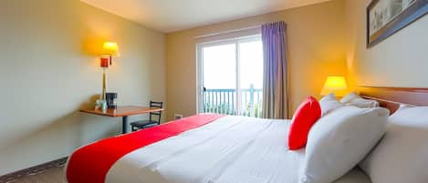 Habitación de hotel con una cama grande, un escritorio con silla, una ventana con cortinas y una lámpara en el escritorio.