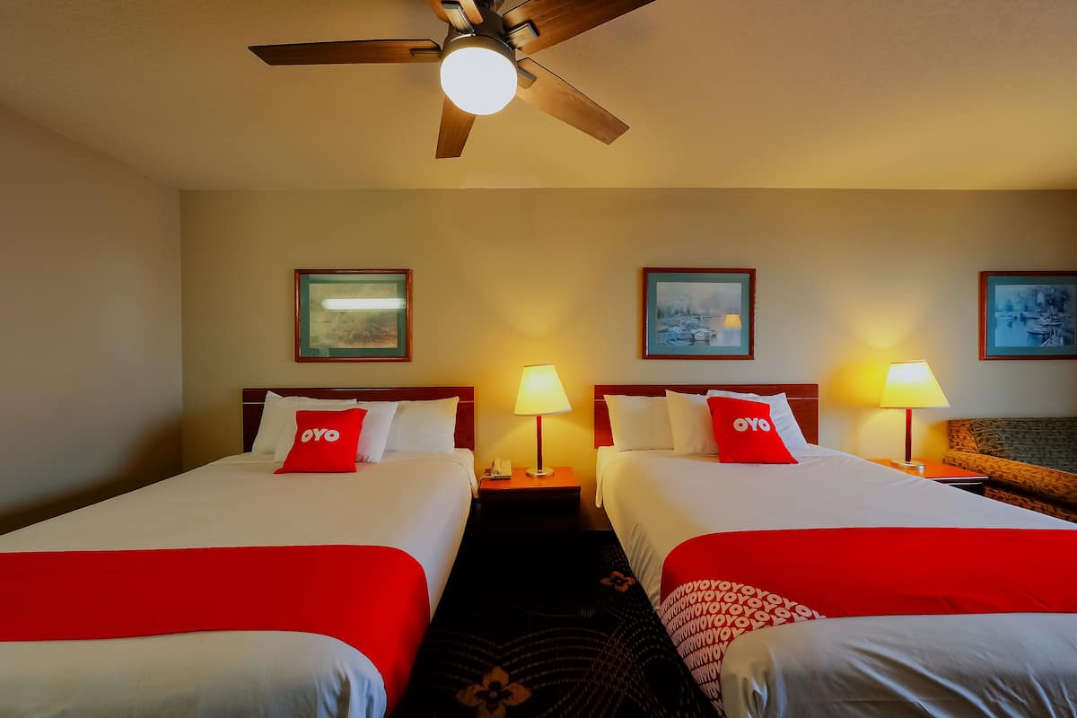 A hotel room with two beds, each with a red OYO pillow, a ceiling fan, and framed pictures on the wall.
