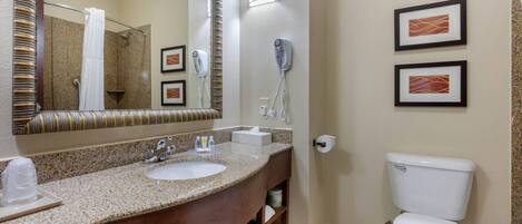 A bathroom with a granite countertop sink, a mirror with a striped frame, a hairdryer, framed pictures on the wall, and a white toilet.