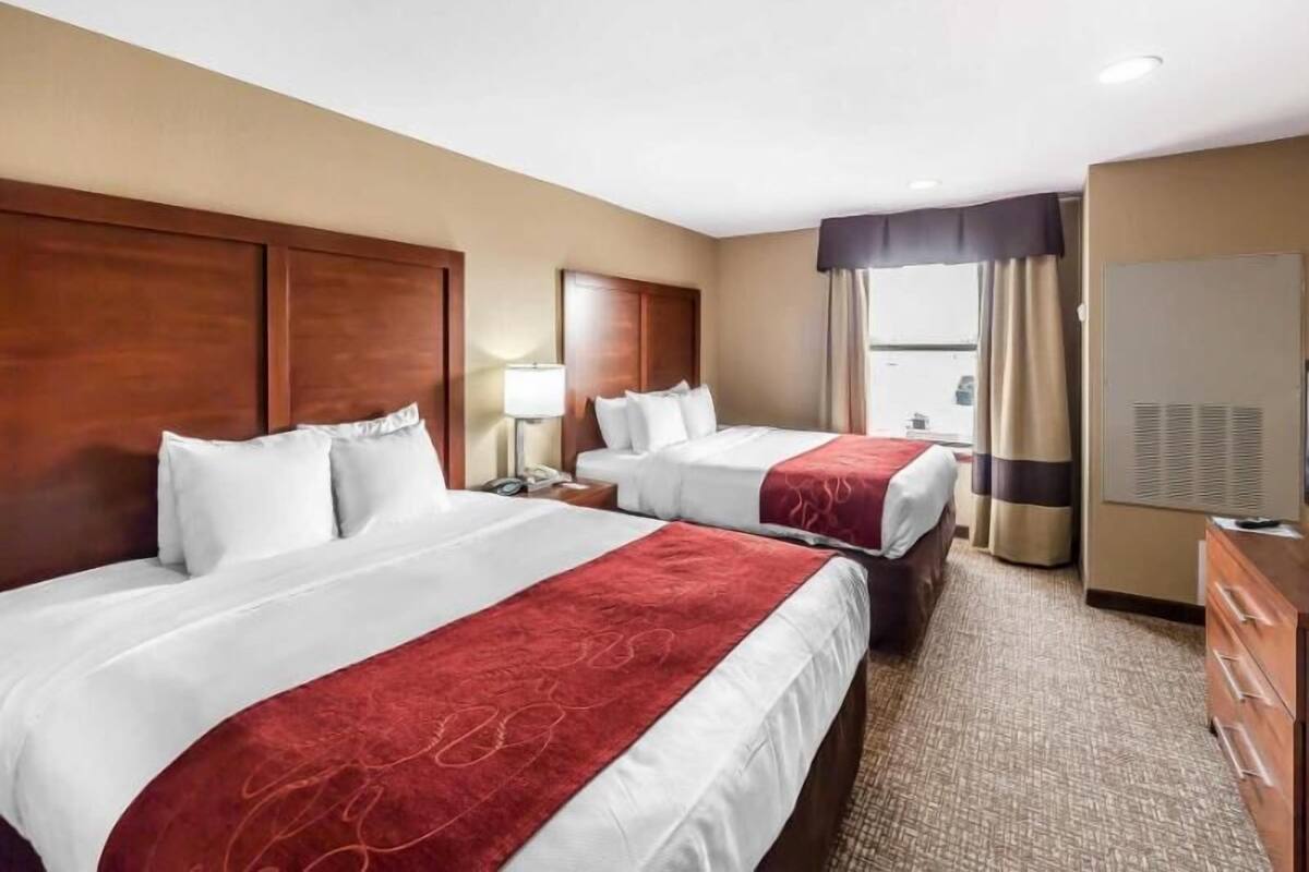 A hotel room with two beds, a wooden headboard, a desk, a lamp, a window with curtains, and an air conditioning unit.