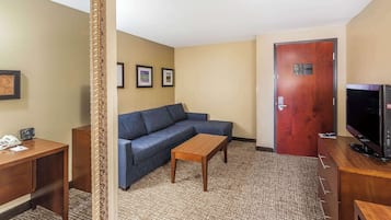 Habitación de hotel con un escritorio de madera, un sofá azul, una mesita de centro de madera, un televisor sobre un mueble de madera y una puerta con una placa.