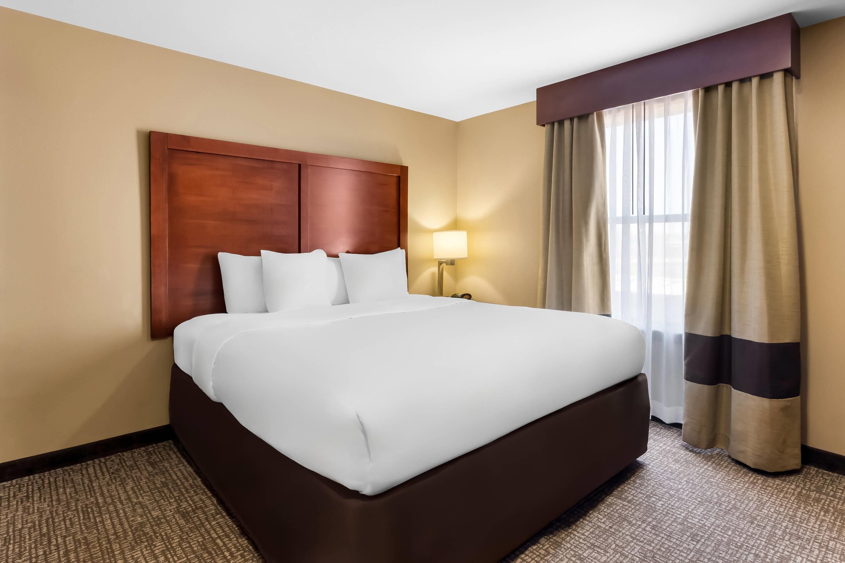 Quarto de hotel com uma cama grande, cabeceira de madeira, mesa de cabeceira com abajur e janela com cortinas.