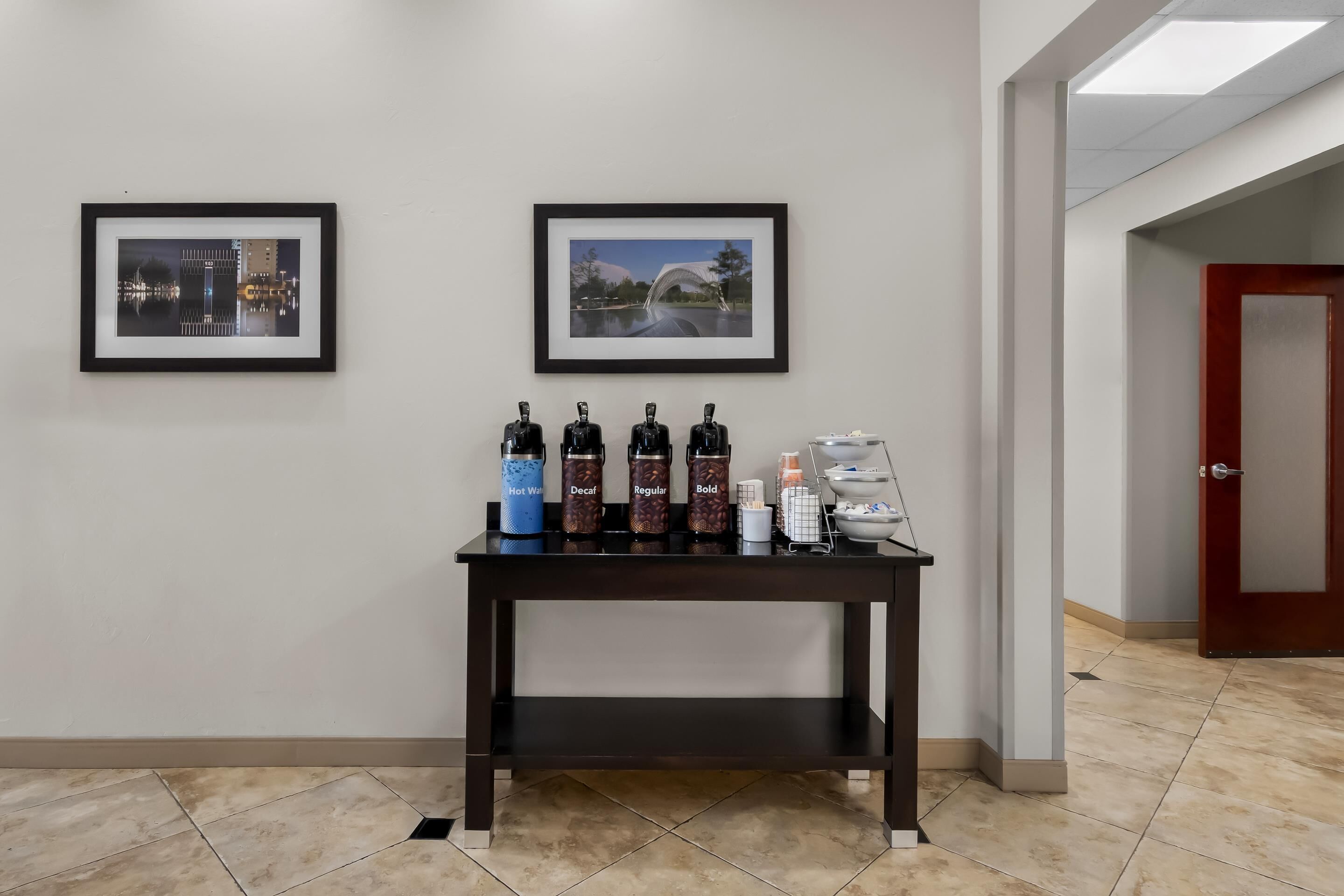 A reception area with a dark wooden table displaying coffee dispensers and a water dispenser. Two framed pictures are hung on the wall above the table.