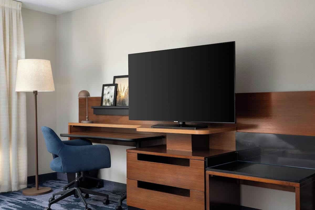 Hotellihuone, jossa on puinen työpöytä, litteänäyttöinen televisio, sininen toimistotuoli, lamppu ja seinällä oleva kehystetty kuva.