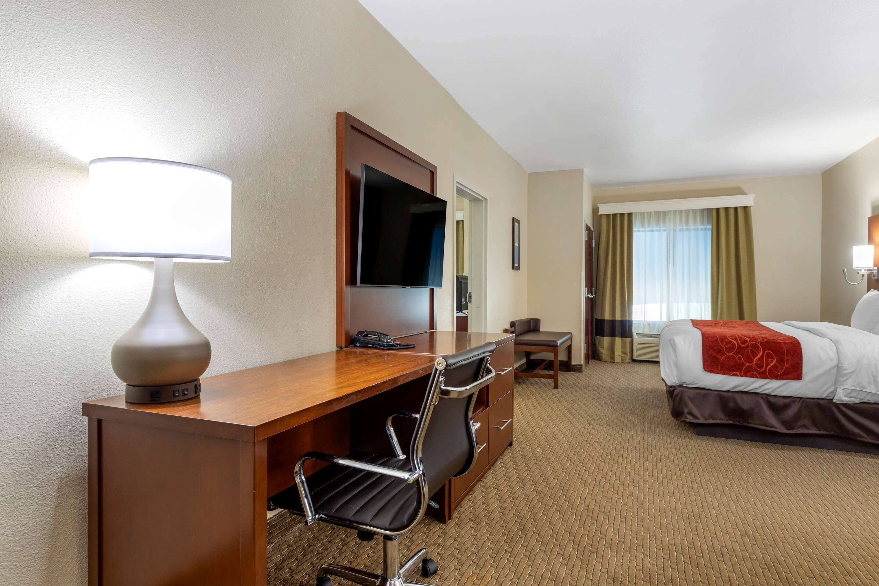 Quarto de hotel com escrivaninha de madeira, uma cadeira, televisão, cama com manta vermelha e janela com cortinas.