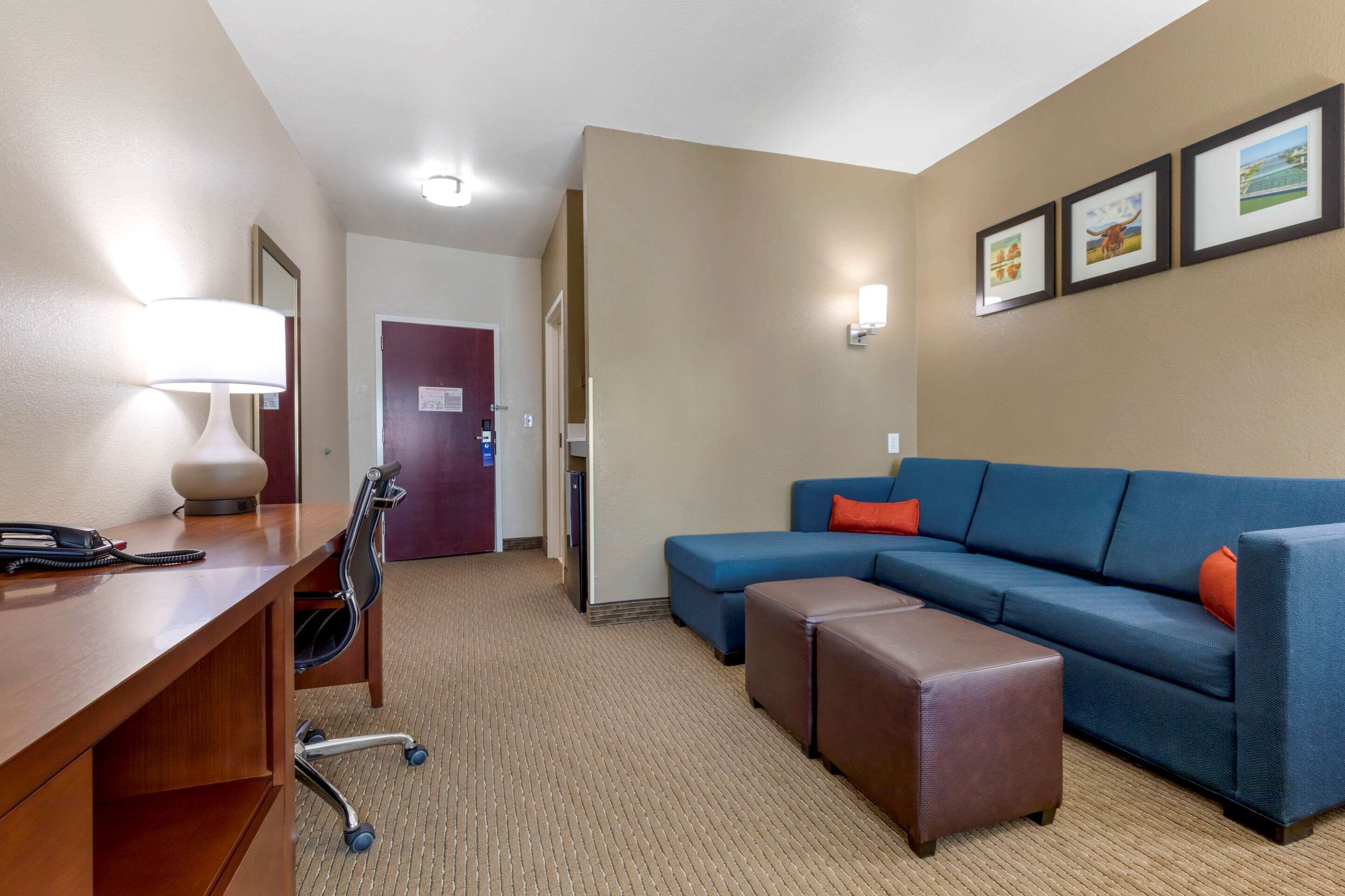 A hotel room with a desk, chair, blue sofa, ottoman, and framed pictures on the wall.
