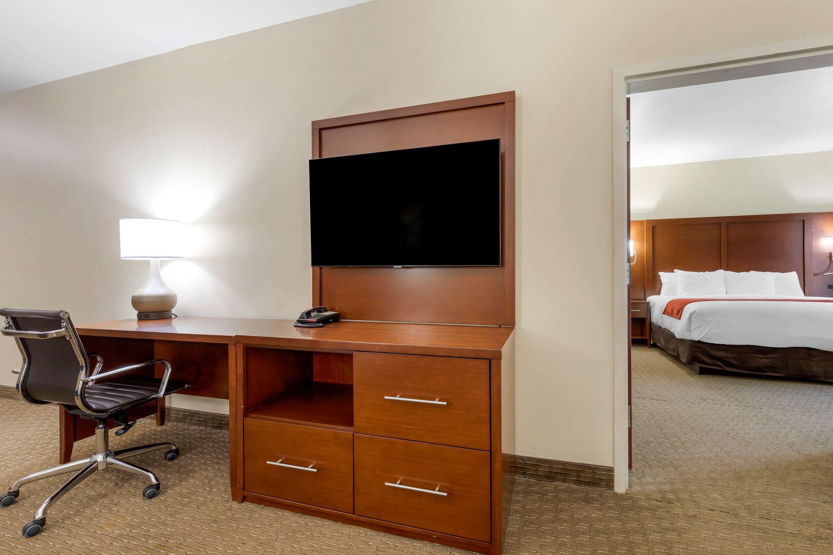A hotel room with a wooden desk, a flat-screen TV, a lamp, a chair, and a bed with white linens.