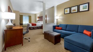 A hotel room with a blue sofa, a desk with a lamp, a bed with a red throw, and a large window with curtains. - Comfort Suites Copperas Cove (Copperas Cove)