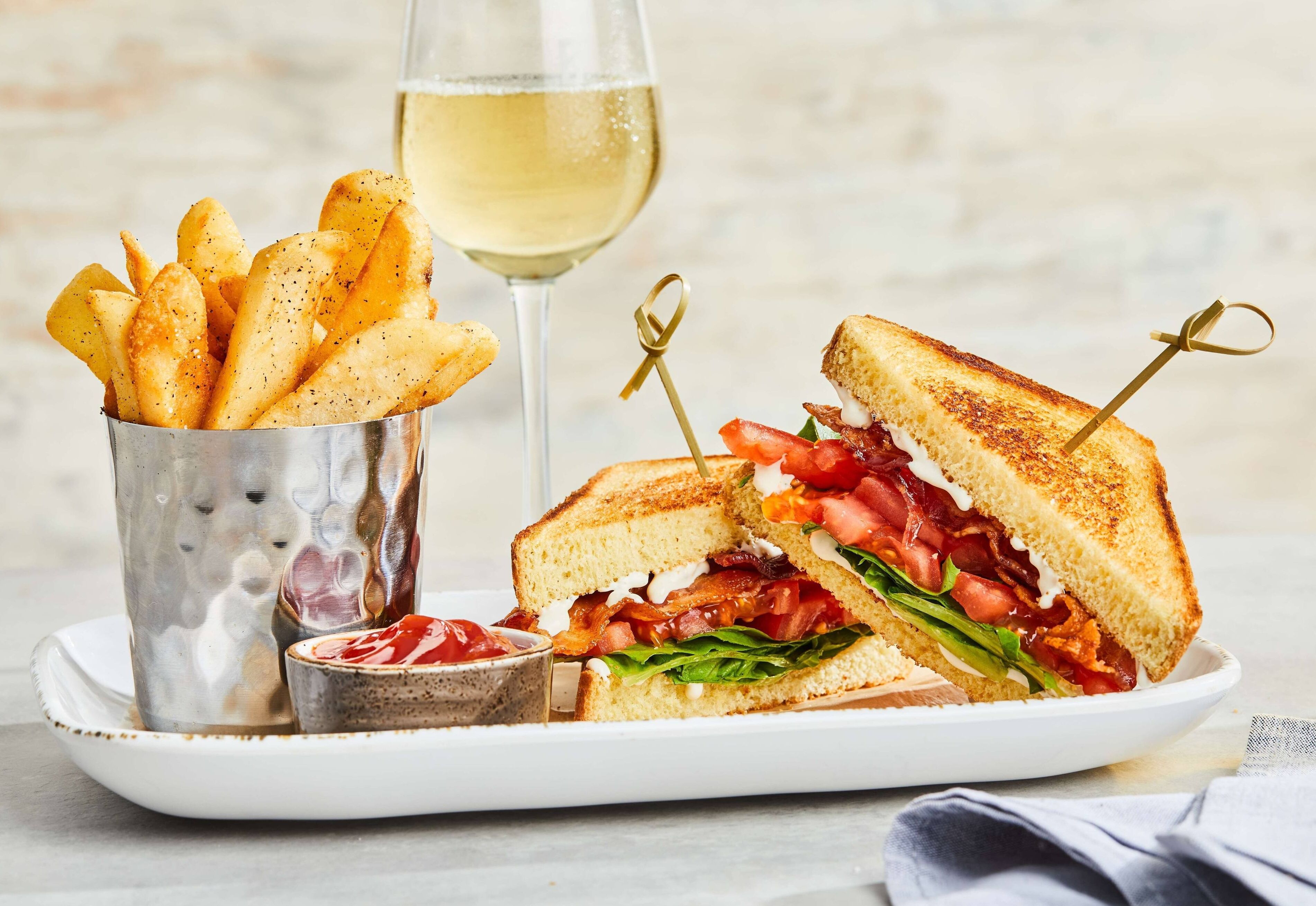 Un plato con un sándwich a la parrilla, una copa de vino blanco y un pequeño recipiente de kétchup.