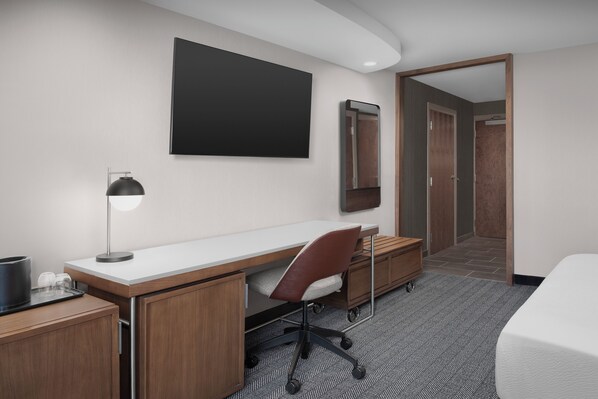 A hotel room with a wooden desk, a chair, a bed, a flat-screen TV, a mirror, and a door leading to another room.