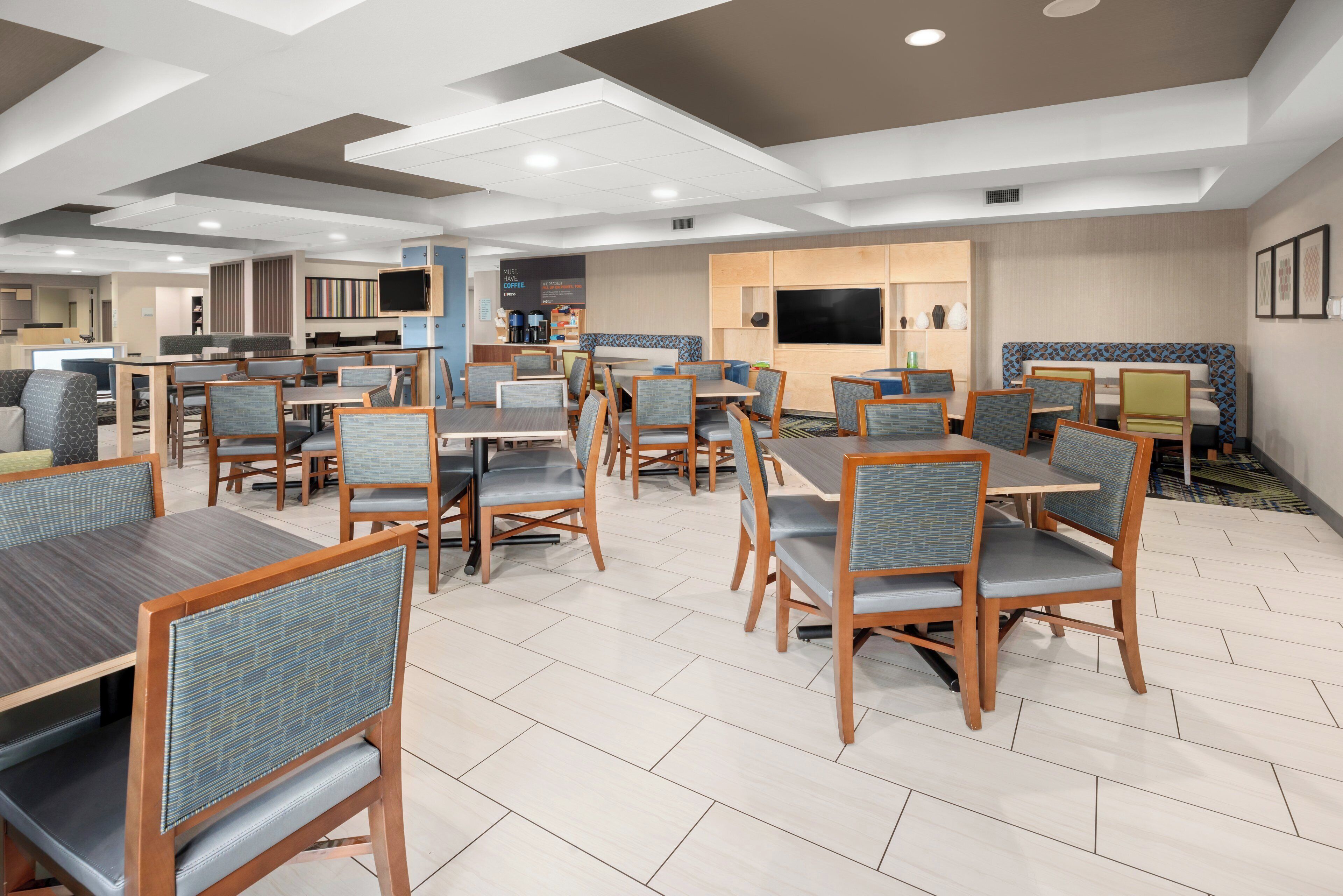 Espaço de refeições de hotel, com várias mesas e cadeiras, um ponto de café e uma tela de televisão.