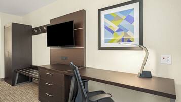 Habitación de hotel con escritorio, televisor de pantalla plana montado en un panel de madera, silla, lámpara de escritorio y un cuadro abstracto enmarcado en la pared.