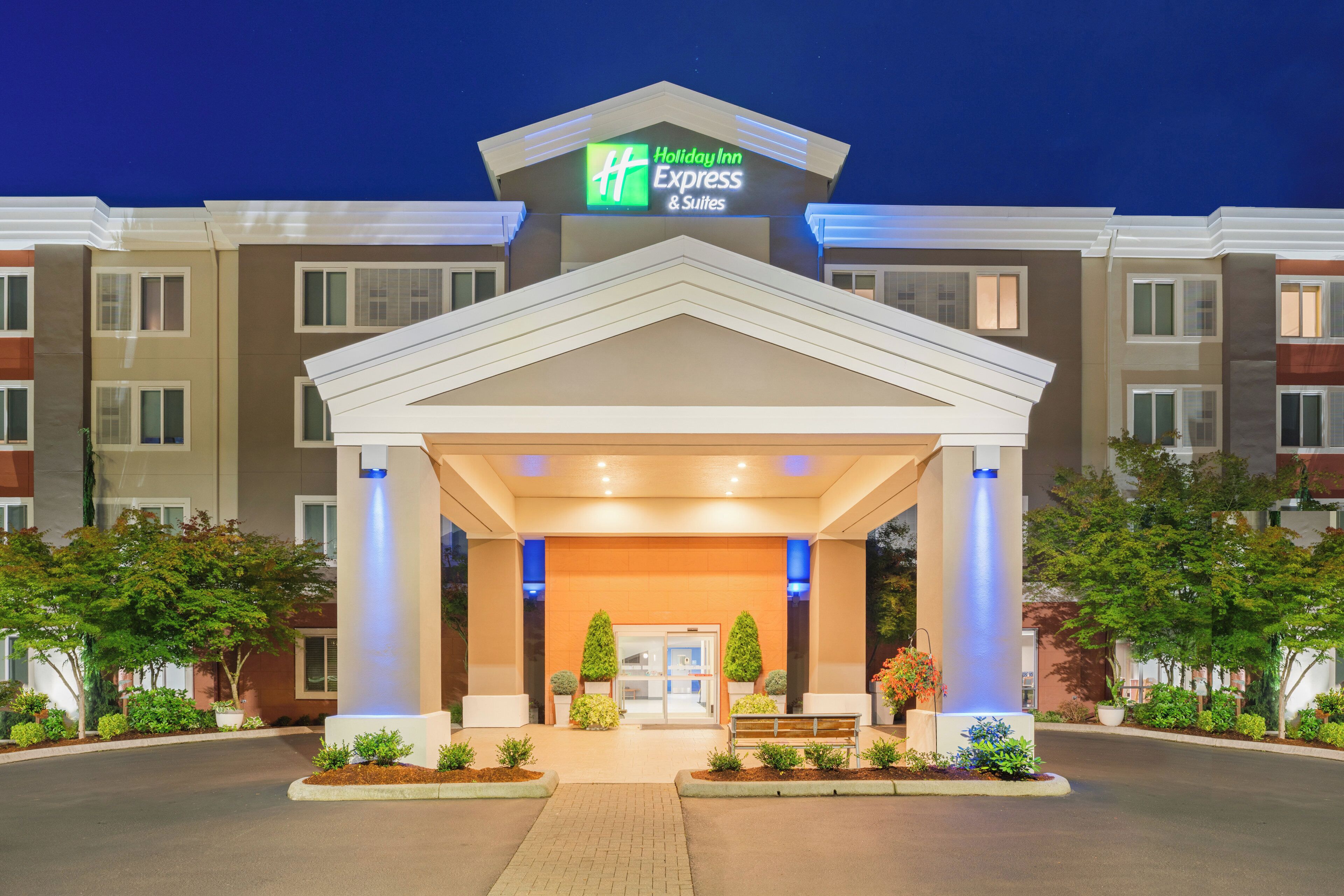 A Holiday Inn Express & Suites building with a covered entrance, potted plants, and a bench.