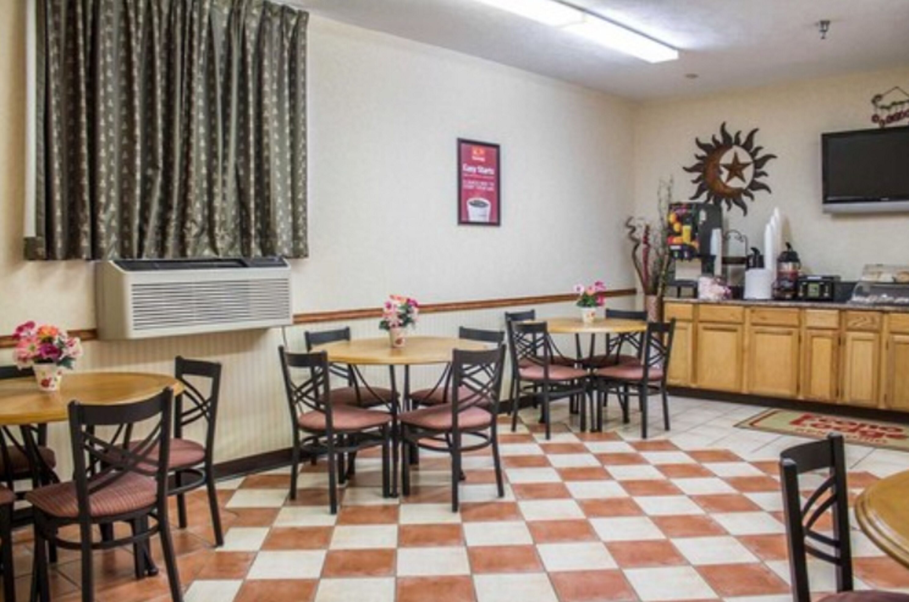 A hotel dining area with checkered flooring, tables with chairs, a wall-mounted TV, and a decorative sun emblem.