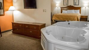 Ein Hotelzimmer mit einer großen, weißen Badewanne, einem hölzernen Schminktisch, einem Bett mit beiger Bettdecke und einem an der Wand befestigten Flachbildfernseher.