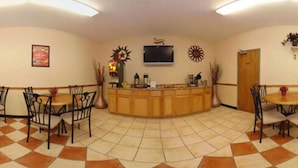 Eine Hotelhalle mit einer Rezeption, zwei Essbereichen mit Tischen und Stühlen, einem an der Wand befestigten Fernseher und einem Fliesenboden.