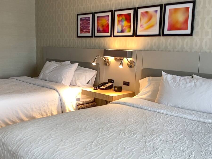 A hotel room with two beds, a nightstand, a lamp, and four framed pictures on the wall.