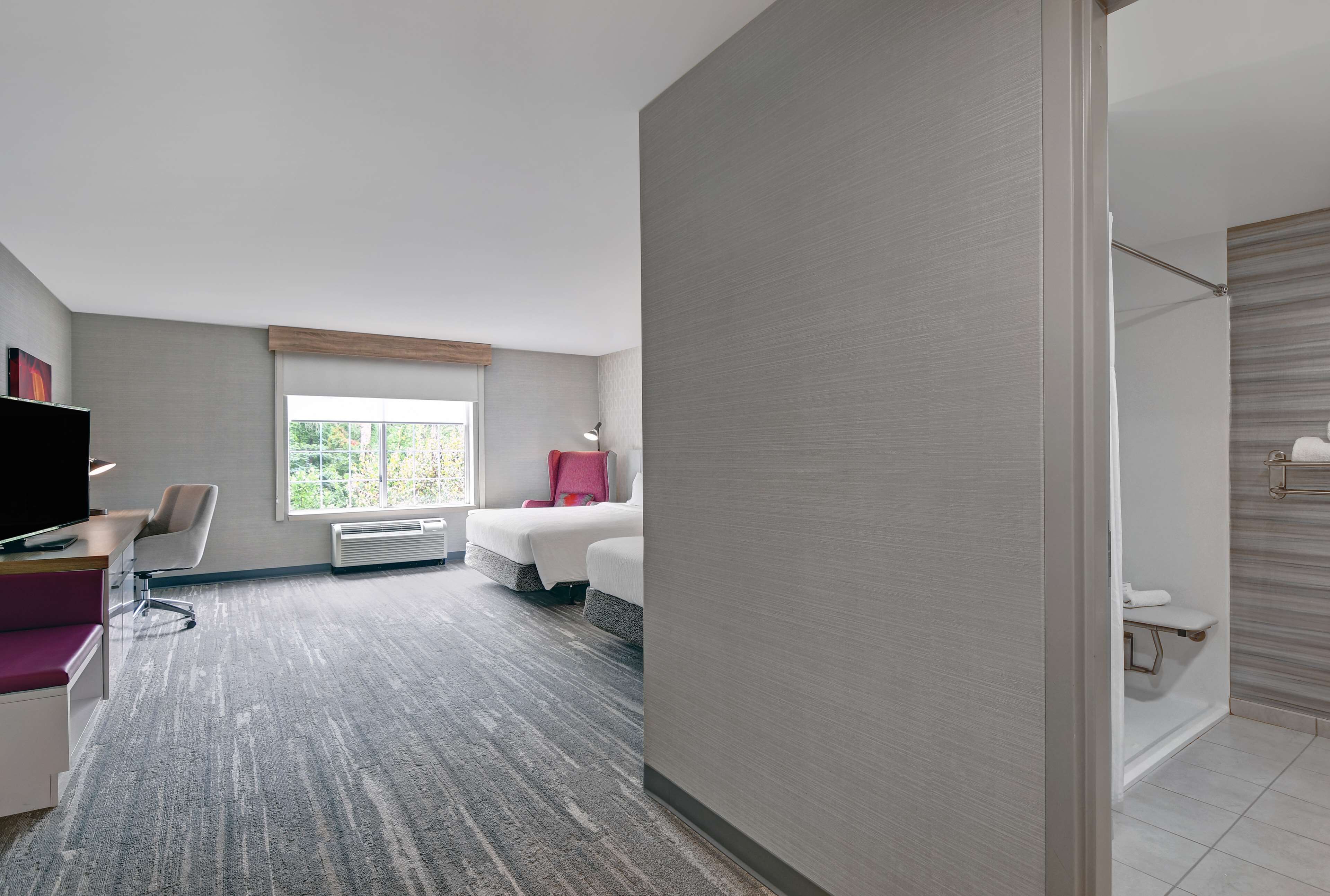 Ein Hotelzimmer mit Schreibtisch, Stuhl, zwei Betten, Fernseher, Fenster und einer Duschkabine.