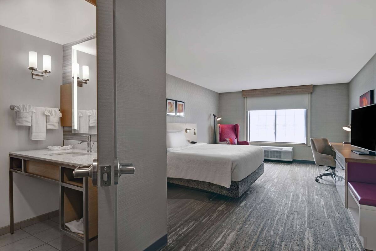 A hotel room with a bed, a desk, a TV, a chair, a bathroom with a sink and mirror, and a window.