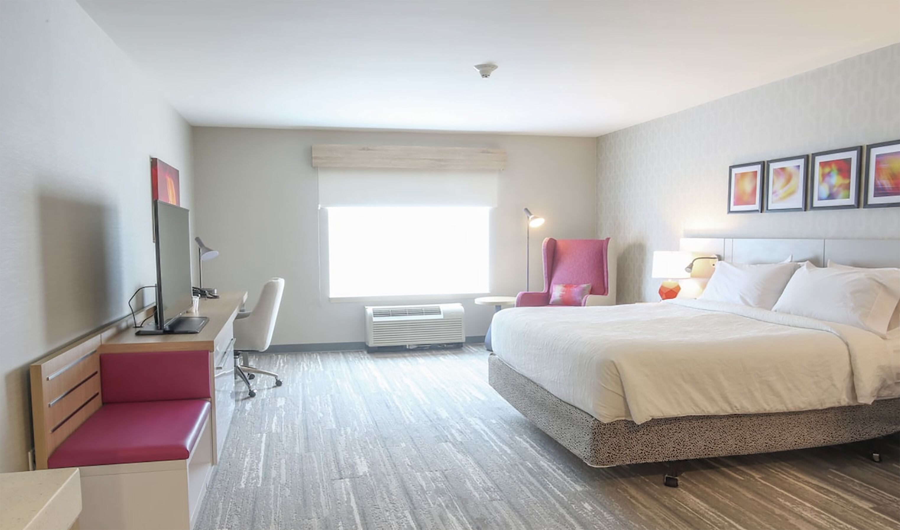 A modern hotel room with a large bed, a desk with a chair, a small table, and a window with blinds.