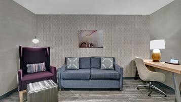Una habitación con un sillón morado, un sofá gris con cojines estampados, un escritorio de madera y una lámpara.