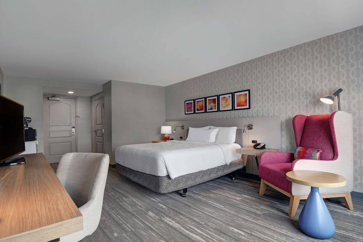 A hotel room with a bed, a desk, a chair, a small table, and a TV.