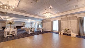 Un amplio salón de baile con varias mesas redondas y sillas, un candelabro y ventanas grandes con cortinas.