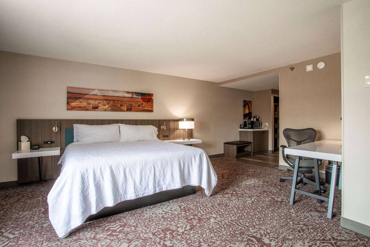 Habitación de hotel con cama, mesita de noche, lámpara, silla, escritorio y un cuadro en la pared.
