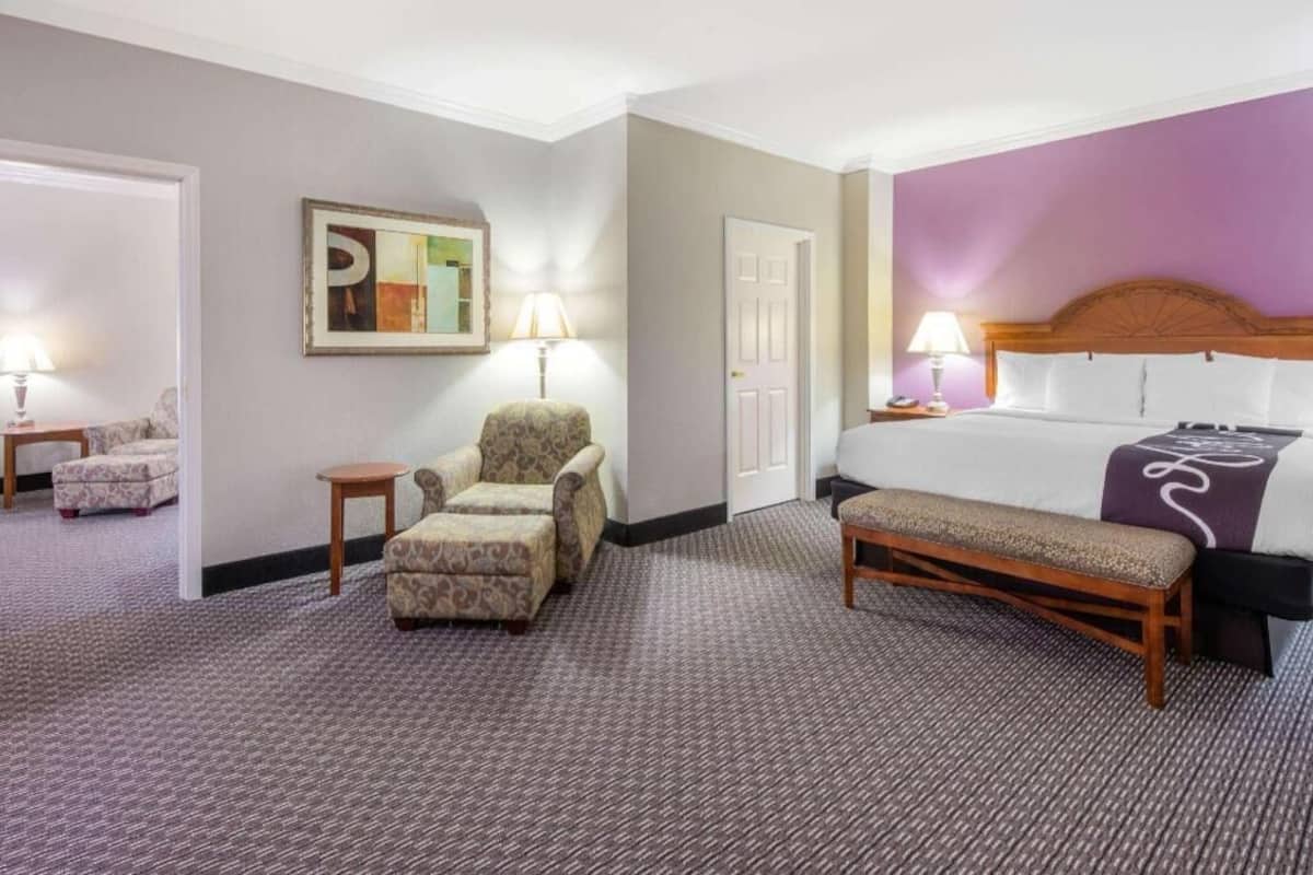 A hotel room with a bed, a chair, a small table, a bench, and a door.