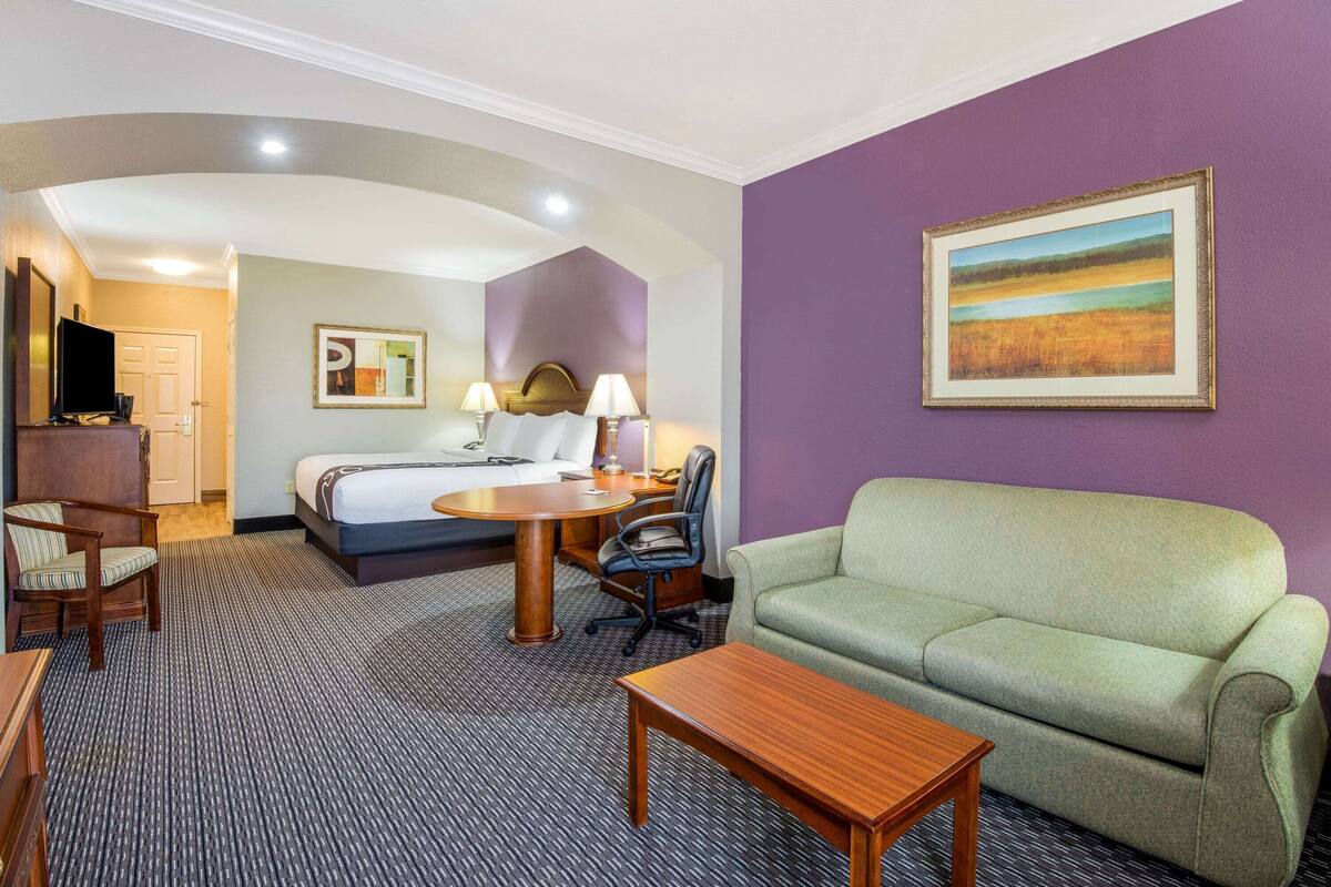 A hotel room with a bed, a green sofa, a wooden table, a desk with a chair, and a TV.
