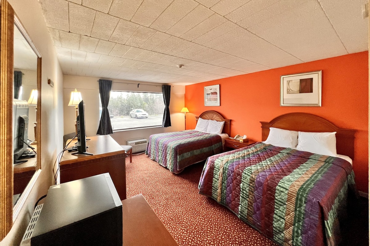 Habitación de hotel con dos camas, un escritorio, una silla, un televisor y ventana con vista a un estacionamiento.