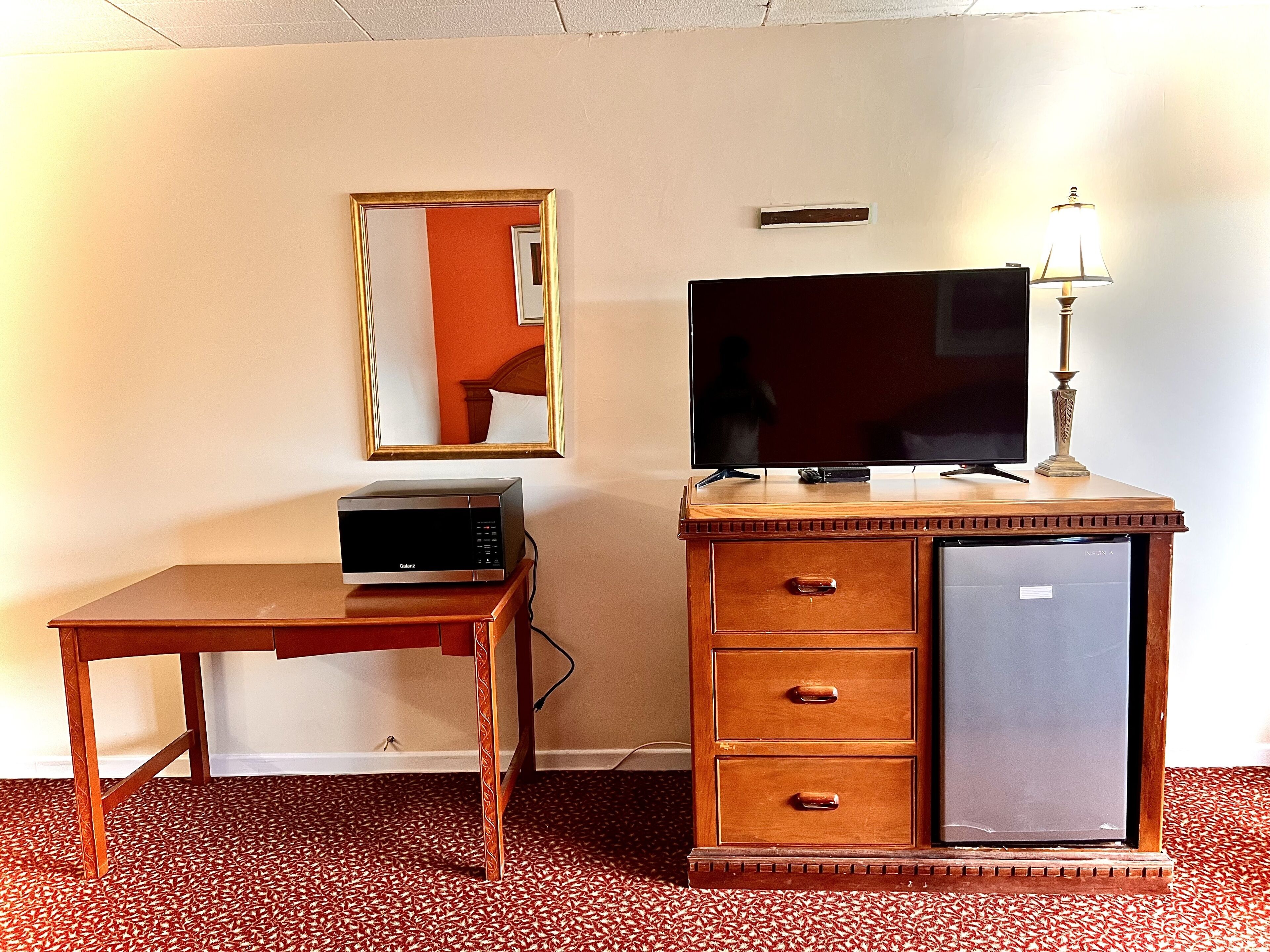 Ein Hotelzimmer mit einem Holztisch, einem Flachbildfernseher, einer Minibar, einer Lampe und einem Spiegel an der Wand.