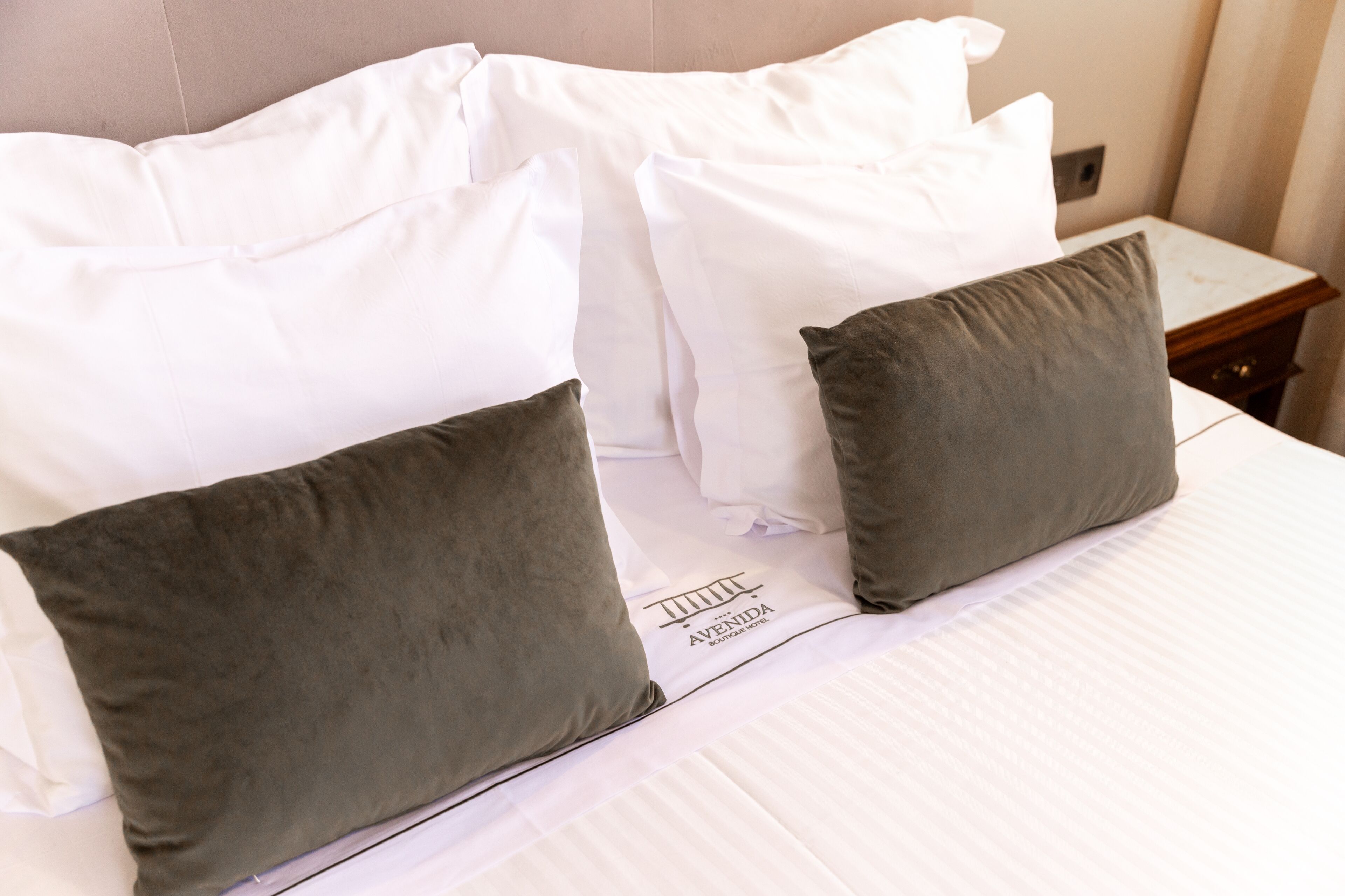 A neatly made bed with white and gray pillows, a clean white bedspread, and a small wooden nightstand with a lamp.