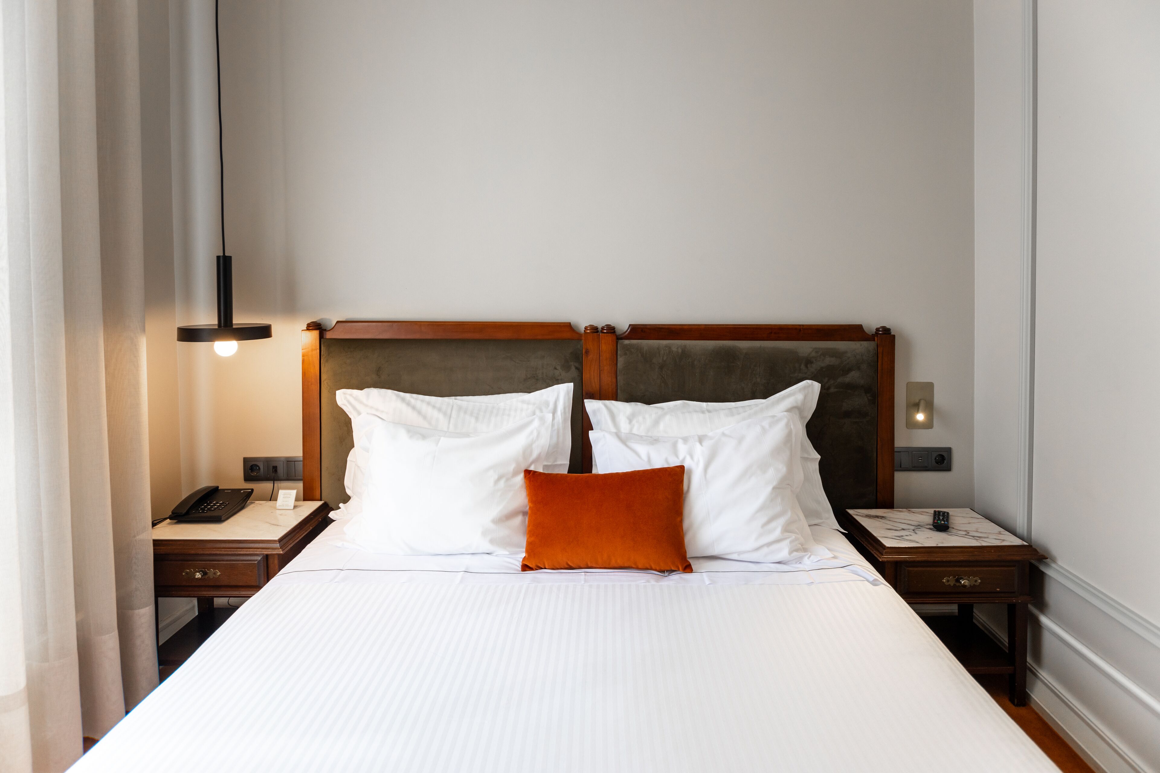Un lit bien fait, avec du linge de lit blanc et un seul oreiller orange, flanqué de deux tables de chevet en bois, sur lesquelles reposent des lampes et un téléphone.