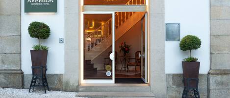 A entrada do Hotel Avenida Boutique, com uma escadaria que leva ao interior, duas plantas em vasos de cada lado e um candelabro visível através da porta de vidro.