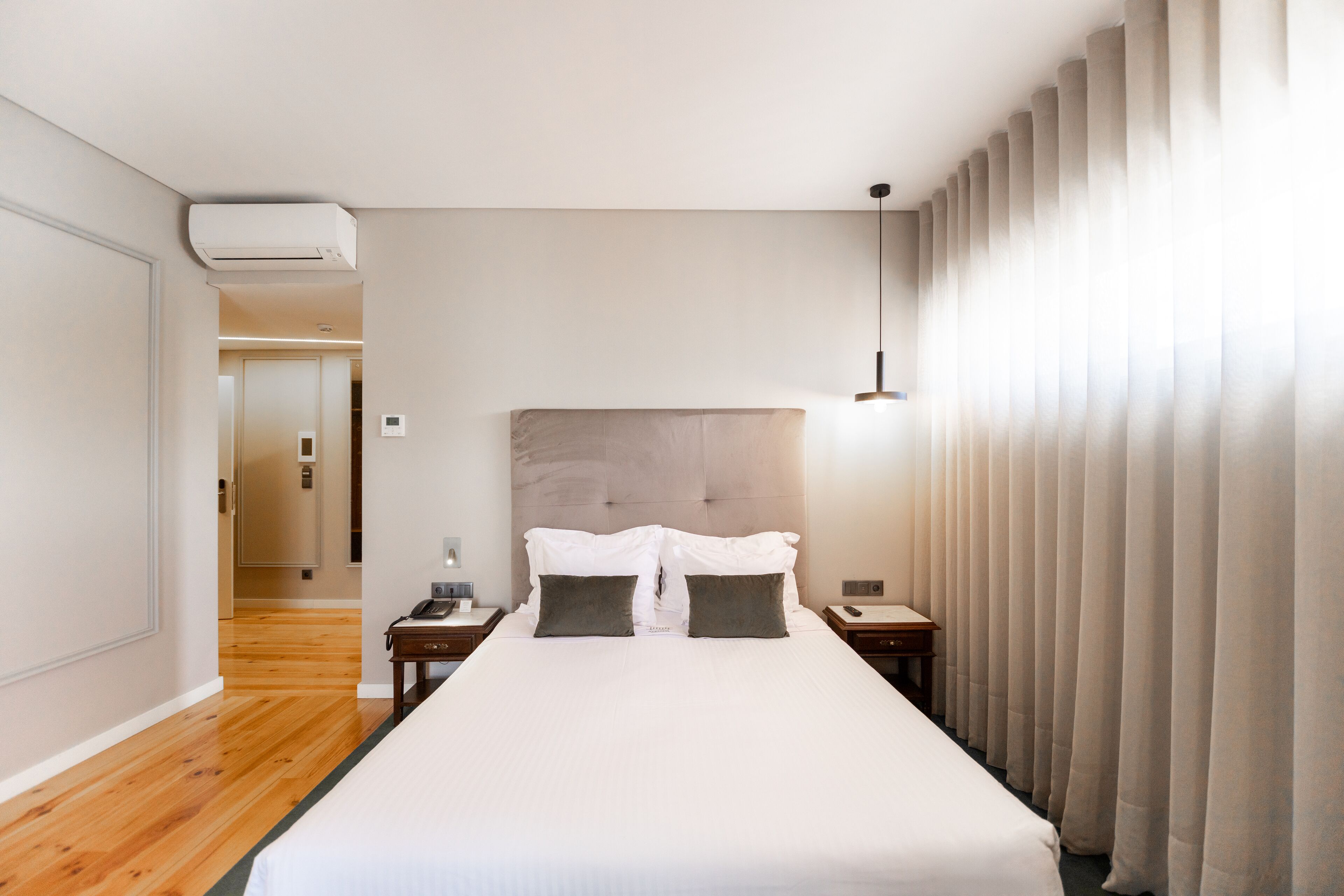 Habitación de hotel con una cama grande, cabecera, mesita de noche con lámpara y ventana con cortinas.