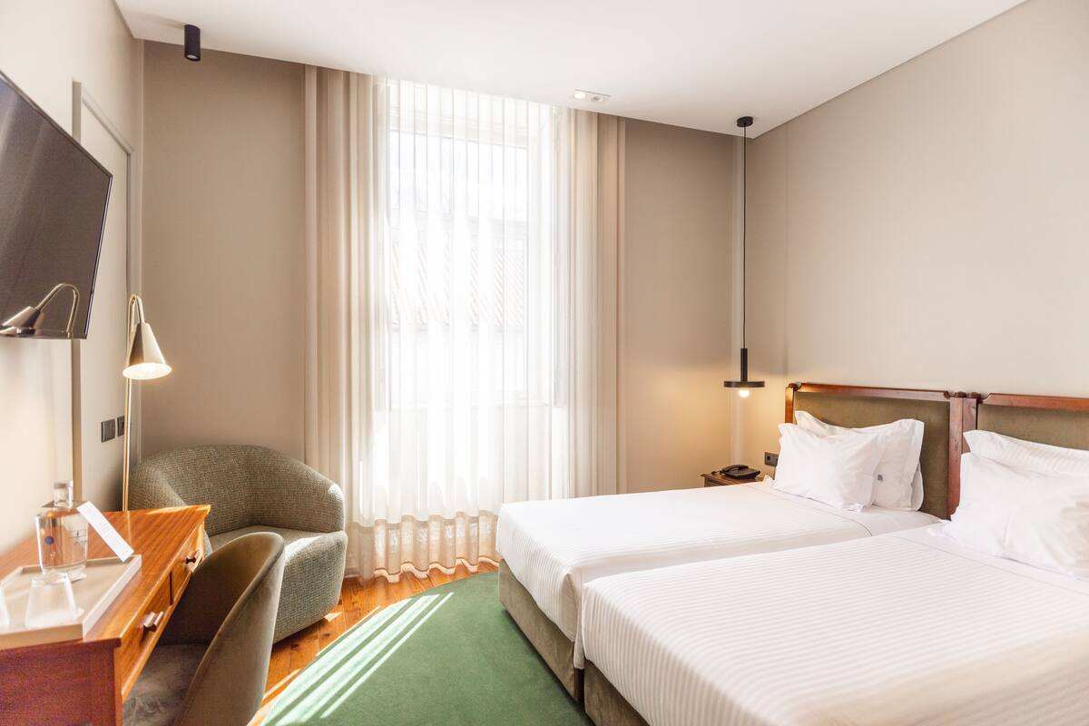 Habitación de hotel con dos camas, un escritorio con silla, televisión y un ventanal con cortinas traslúcidas.