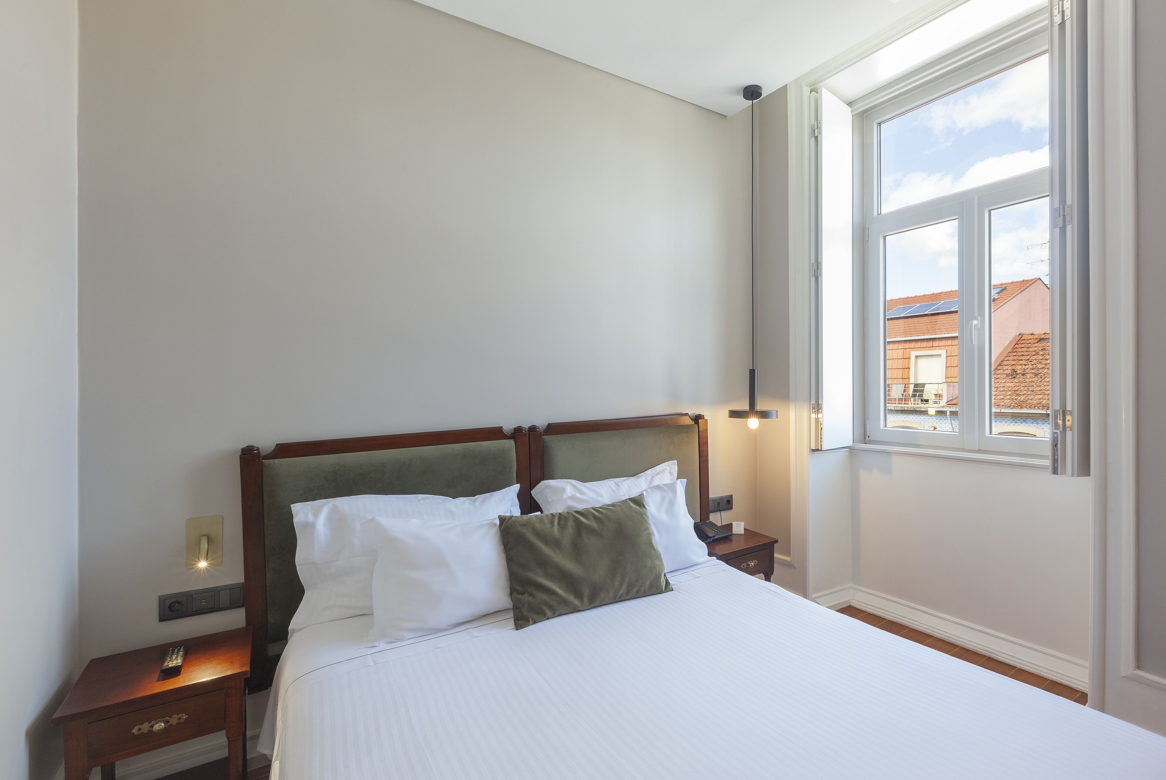 A neatly made bed with white linens and a green pillow, a wooden headboard, a bedside table with a remote control, and a window with a view of buildings outside.