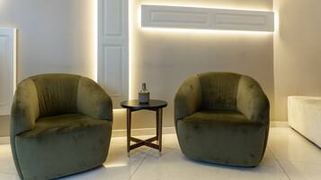Dos sillones verdes, una mesita redonda con un florero, y un sofá blanco en una habitación moderna, bien iluminada y de diseño minimalista y ordenado.