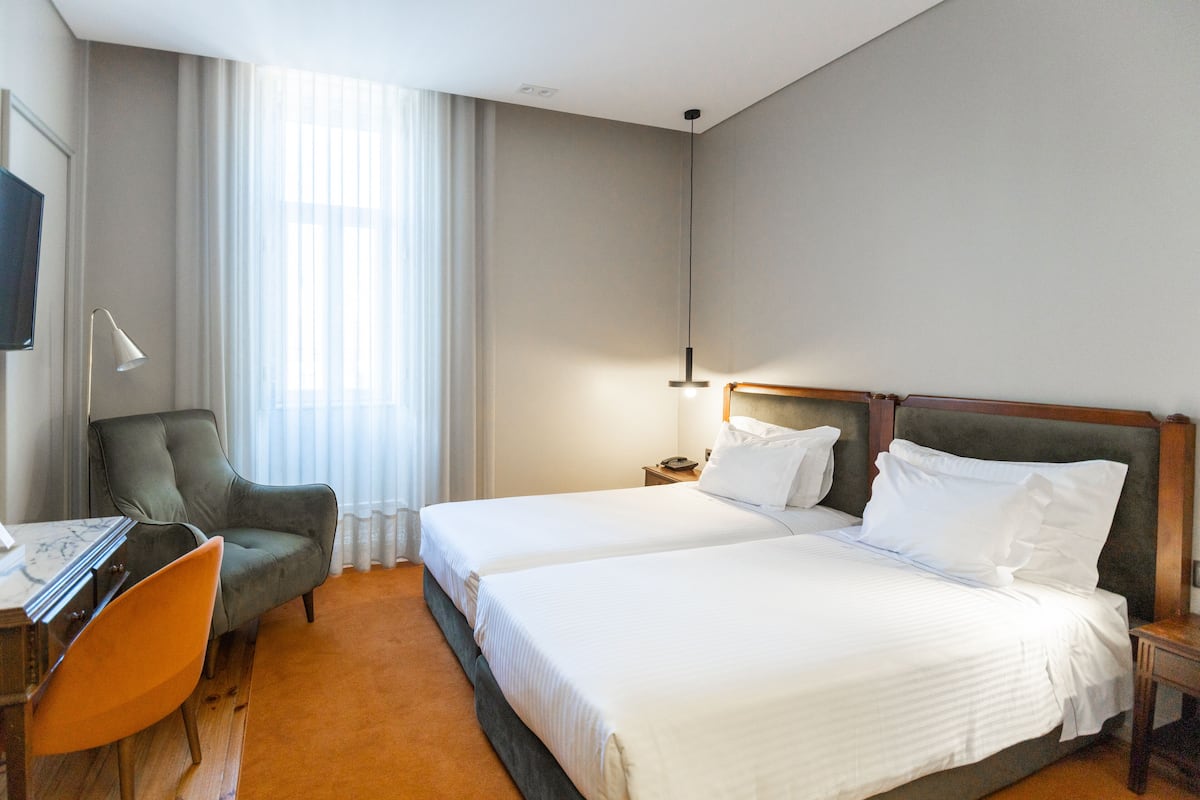 Habitación de hotel con dos camas, un escritorio con silla, televisor y un ventanal con cortinas.