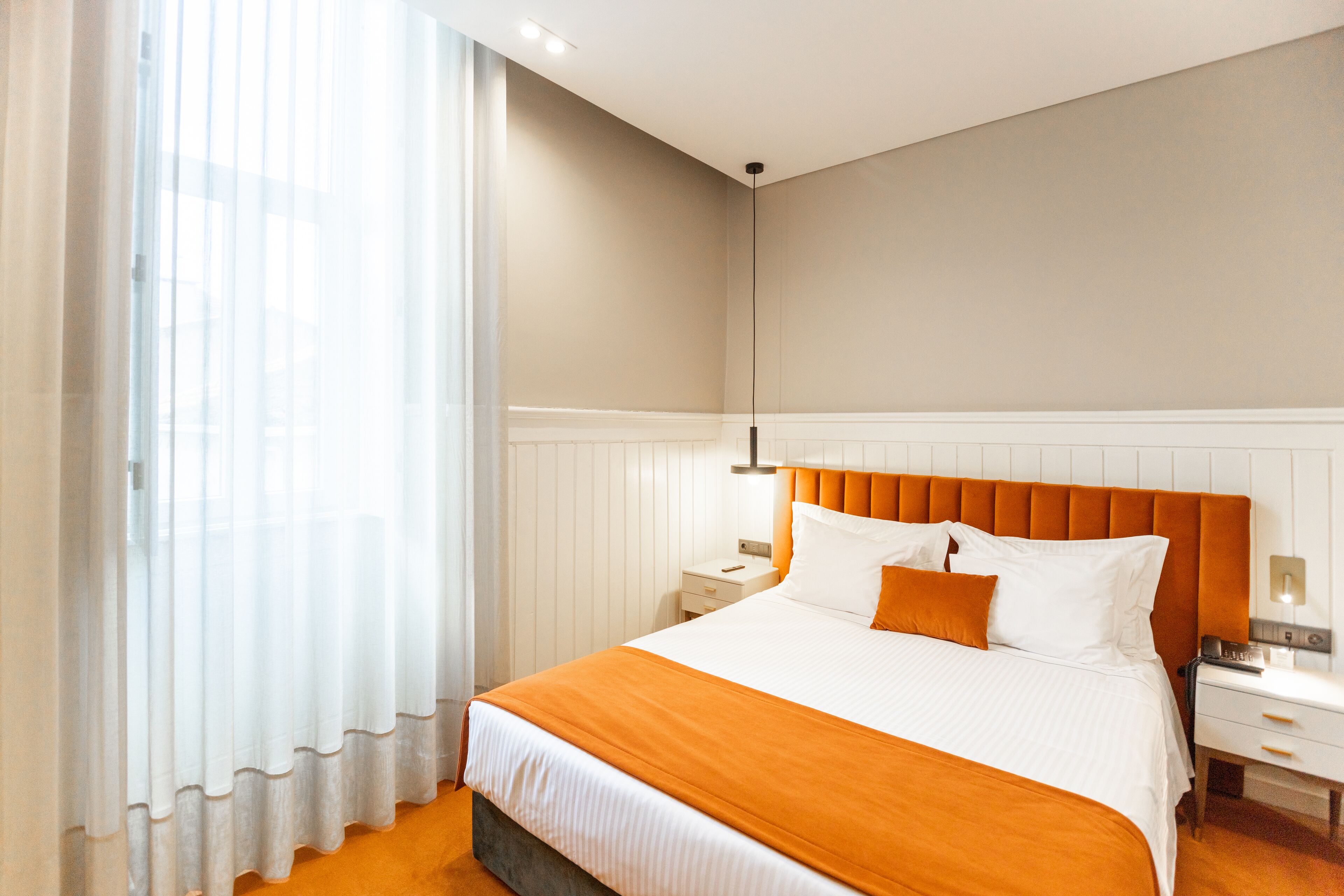 A hotel room with a large bed, a nightstand, a lamp, and a window with sheer curtains.