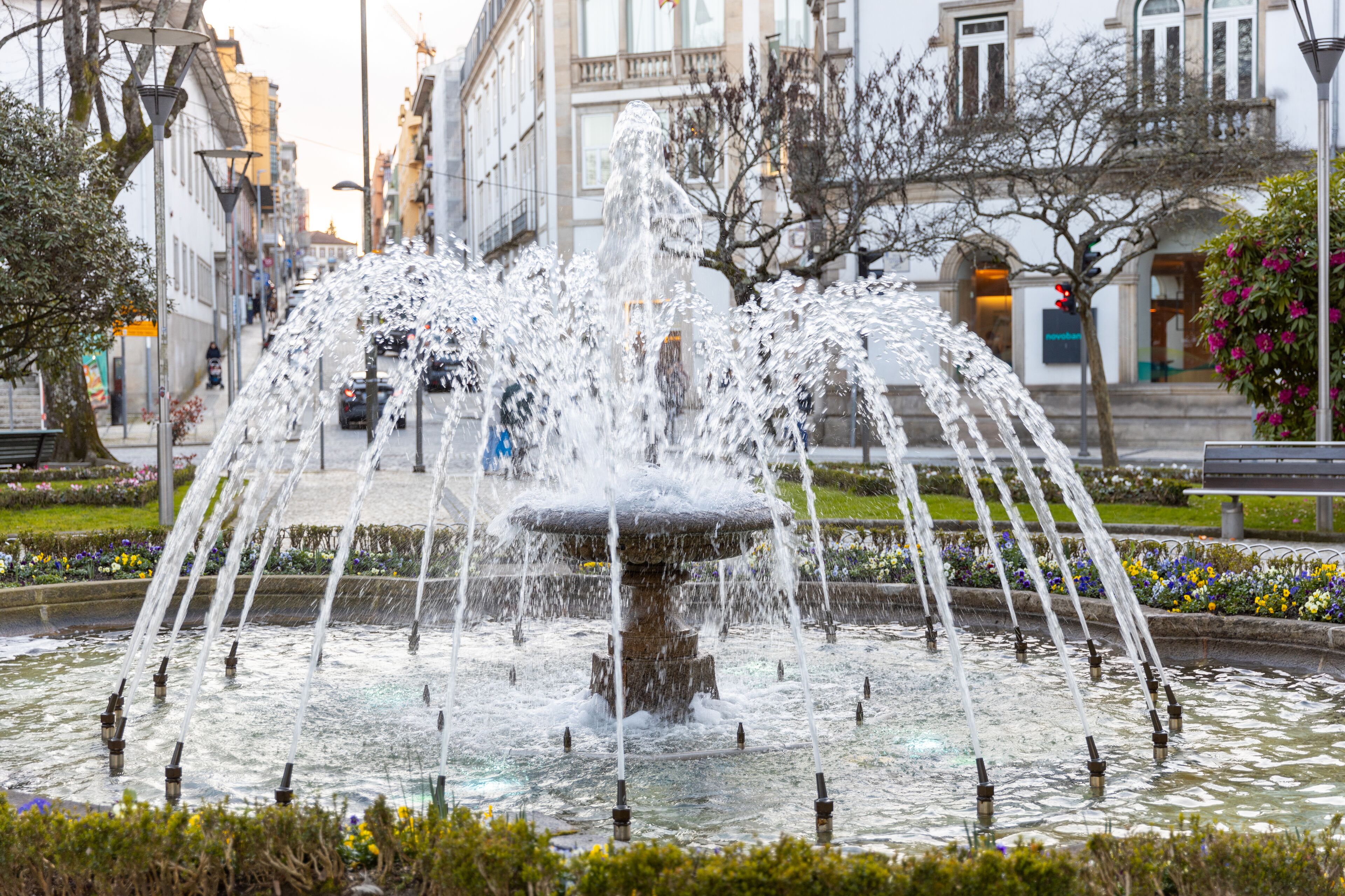 A fountain with multiple water jets in a public square with buildings and benches around.