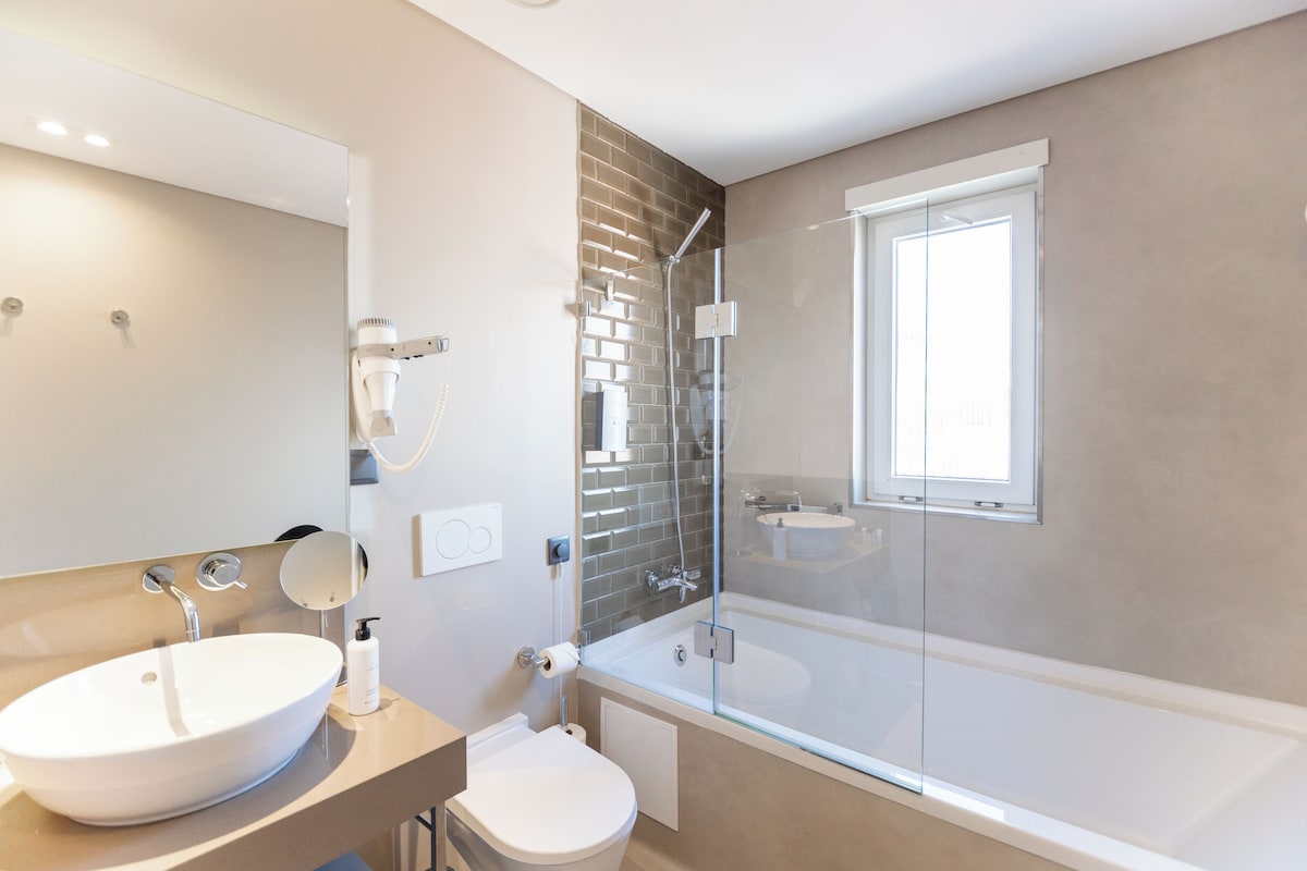 A modern bathroom with a large white sink, a toilet, a glass-enclosed shower, and a window.
